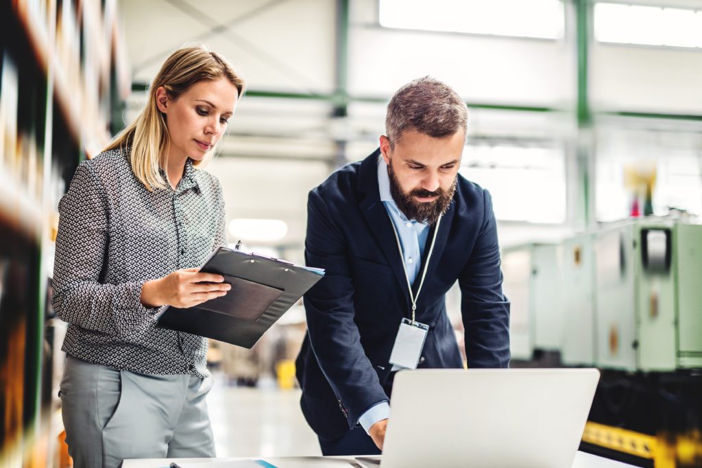 A portrait of a serious mature industrial man and woman engineer with laptop in a factory, working.